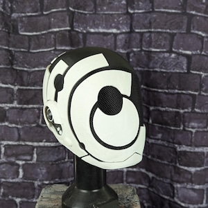 May include: A black and white futuristic helmet, presented on a black stand. The helmet has a mesh visor and white geometric patterns. The backdrop is a dark brick wall.