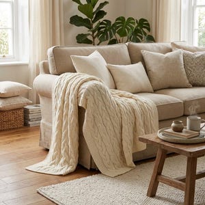 May include: A cream-colored cable knit throw blanket draped over a beige sofa. The sofa features several matching cream and beige throw pillows. The blanket has a thick, textured cable pattern and hangs down the side of the sofa seat.