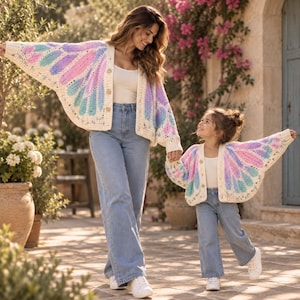 May include: Two matching crochet cardigans featuring a cream base with a vibrant wing-like pattern in shades of pink, purple, and teal. Each cardigan has a button-front closure, long sleeves, and a loose fit, worn with white tops and blue jeans.