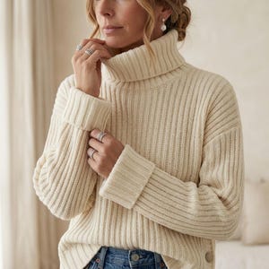 May include: A cream-colored, ribbed knit sweater featuring a high turtleneck collar and long sleeves with folded cuffs. The sweater is worn with blue denim jeans. The design includes a vertical ribbed texture throughout the garment.