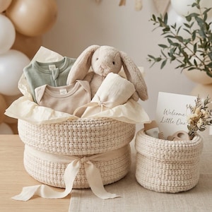 Peut inclure: Un grand panier crocheté couleur crème contenant un body vert sauge, un body beige, une peluche lapin beige et une couverture crème pliée. Un plus petit panier crocheté assorti contient une carte avec le texte Welcome Little One.