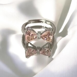 Può includere: Un anello d'argento con una pietra preziosa rosa a forma di fiocco. L'anello è incastonato con pietre preziose trasparenti più piccole intorno al fiocco.