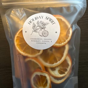 Może przedstawiać: Przezroczysta plastikowa torba wypełniona suszonymi plasterkami pomarańczy, laskami cynamonu i okrągłą etykietą. Na etykiecie widnieje napis "HOLIDAY SPIRIT" oraz lista żurawiny, pomarańczy, rozmarynu, goździków i cynamonu. Plasterki pomarańczy są w żywym pomarańczowym kolorze.