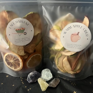 Puede incluir: Dos bolsas transparentes de rodajas de fruta deshidratada, etiquetadas como "Fall Harvest" y "Warm Apple Cider". La bolsa "Fall Harvest" incluye manzanas, naranjas, anís estrellado, hojas de laurel, clavos y canela. La bolsa "Warm Apple Cider" contiene manzanas, canela, vainilla, pimienta de Jamaica, clavos y nuez moscada. Varios cristales coloridos en la parte inferior.