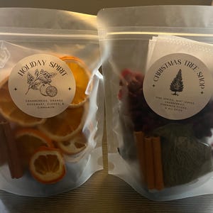 Puede incluir: Dos bolsas transparentes llenas de popurrí navideño. Una bolsa presenta rodajas de naranja secas, palitos de canela y una etiqueta que dice "Holiday Spirit". La otra bolsa contiene arándanos, ramitas de pino y una etiqueta que dice "Christmas Tree Shop".