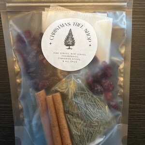 May include: A clear plastic bag containing Christmas-themed ingredients. The bag includes cinnamon sticks, pine sprigs, cranberries, mint leaves, and allspice. A circular label reads "Christmas Tree Shop".