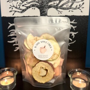 Può includere: Un sacchetto trasparente pieno di fette di mela essiccate, etichettato "Warm Apple Cider". L'etichetta include un disegno di tazza ed elenca gli ingredienti. Due lumini accesi affiancano il sacchetto.