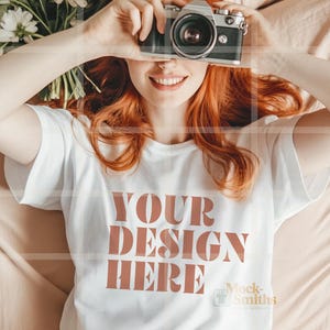 May include: A white t-shirt with the text "YOUR DESIGN HERE" printed in brown on the front. The shirt is being worn by a person with red hair who is holding a camera up to their face.