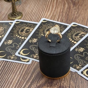 May include: A gold ring with a clear, faceted gemstone is displayed on a black ring holder. Tarot cards with a celestial design are arranged around the ring. The cards feature moons, suns, and floral elements.