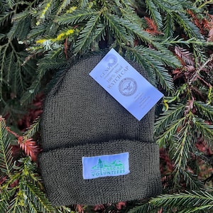 May include: A dark green wool watch cap with a white patch that says "CASCADE VOLUNTEERS" in green letters. The cap is sitting on a bed of green pine needles. A white tag is attached to the cap that says "GENUINE GOVERNMENT ISSUE 100% Wool WATCH CAP" with a seal of the Department of the Navy.