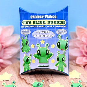 May include: A package of "Sticker Flakes" featuring "Tiny Alien Buddies" stickers. The package includes 10 alien stickers, 5 star stickers, and 5 different designs. The stickers are green aliens with large eyes and yellow stars. The package is on a wooden surface.