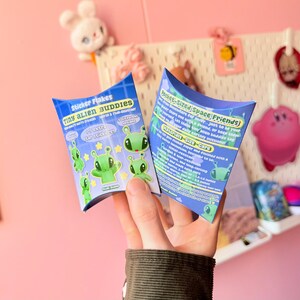 Rear view of the pastel blue pillow box packaging held upright against a pink background with large artificial flowers. The back panel displays printed product details in colorful blocks of text, including quantity and material information. The compact size of the box is visible in proportion to the hand holding it, reinforcing its gift-ready format and structured shape.