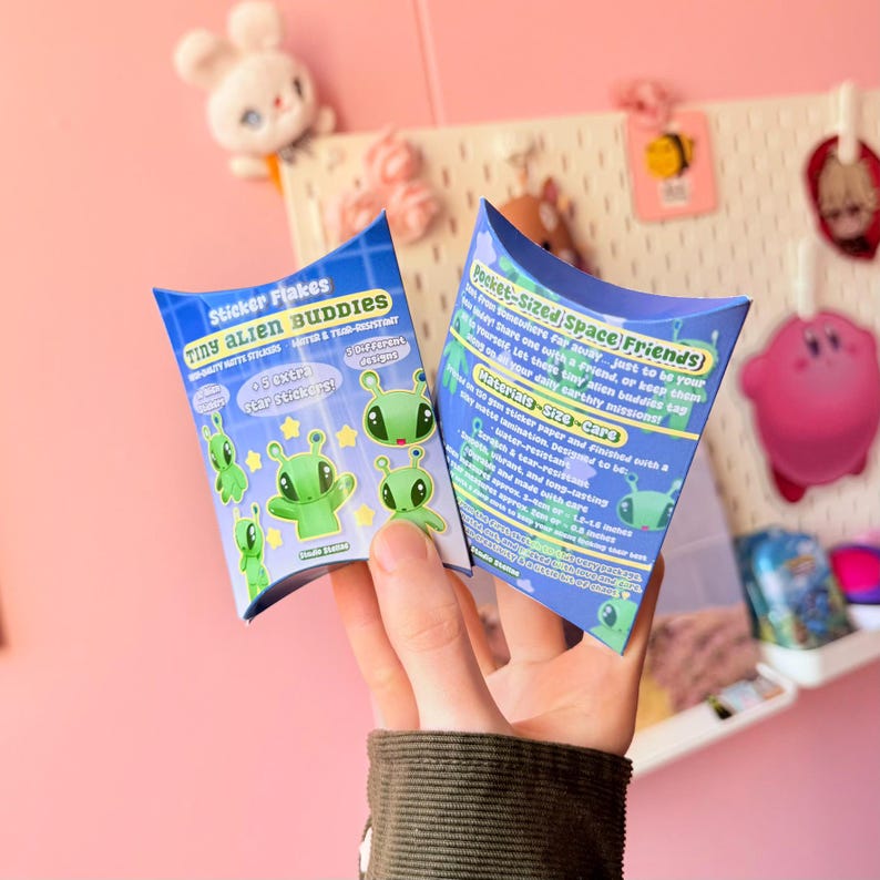 Rear view of the pastel blue pillow box packaging held upright against a pink background with large artificial flowers. The back panel displays printed product details in colorful blocks of text, including quantity and material information. The compact size of the box is visible in proportion to the hand holding it, reinforcing its gift-ready format and structured shape.