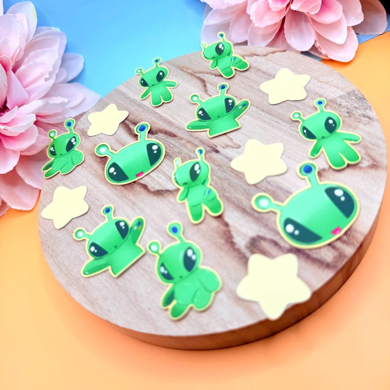 Full overhead shot of the alien and star sticker flakes arranged evenly on a circular wooden board. The green alien characters vary in pose and facial expression, each outlined in bright borders, while smaller yellow star stickers fill the gaps. The pastel background and floral props frame the arrangement, clearly showing the complete 15-piece set and scale consistency.