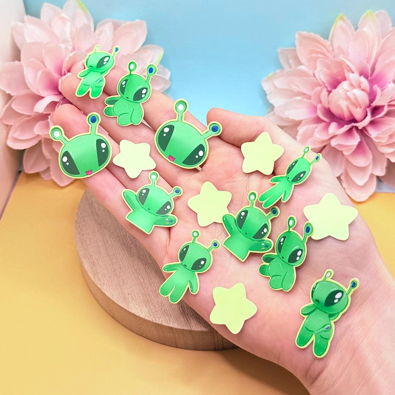 Close-up of a hand resting on a round wooden display board, with several green alien sticker flakes placed across the palm and fingers. Small yellow star stickers are arranged between them. The tray sits on a soft yellow surface with pink flowers framing the scene. The image clearly shows the smooth matte lamination, die-cut edges, and relative scale of the 2–4 cm stickers against the hand.