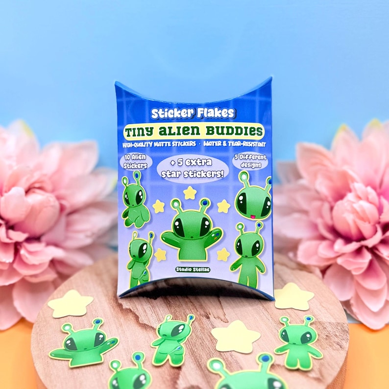 Pastel blue pillow box labeled “Sticker Flakes – Set of 15 Alien Stickers” standing upright against a pink and yellow background with large blush flowers on both sides. In front of the box, green alien character sticker flakes and small yellow star stickers are arranged neatly on the surface. Each sticker has a bold colored border and matte finish. The full 15-piece set is visible, showing size variation between the 3–4 cm aliens and smaller star accents.