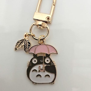 以下が含まれることがあります： ピンクの傘を持った漫画のキャラクターが特徴の、金色の金属製キーホルダー。キャラクターは黒、白、グレーです。小さな金の葉のチャームがキャラクターの隣にぶら下がっています。キーホルダーには金の留め金が付いています。