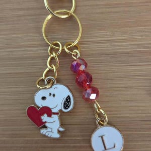 May include: A gold-toned metal keychain featuring a lobster clasp, a white enamel charm of Snoopy holding a red heart, a strand of three faceted red beads, and a small circular white enamel charm with the black capital letter L printed on it.