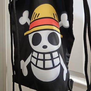 May include: Black drawstring bag with a white skull and crossbones design, a yellow hat and a red band. The bag has black drawstrings and is likely made of a durable fabric, suitable for carrying items.