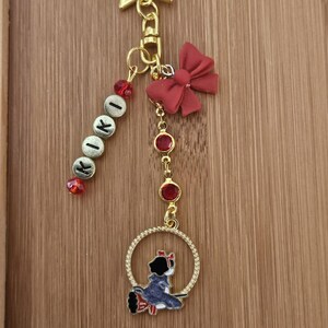 May include: Gold-toned keychain with a star-shaped clasp, featuring a red bow, red gem accents, and a charm of a cartoon character in a circle. The keychain also includes a beaded strand spelling "KOKI".
