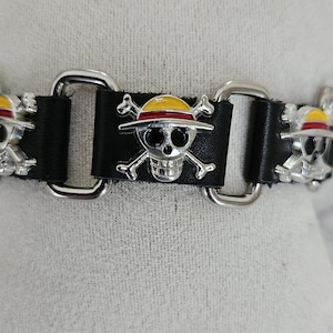 May include: Black leather bracelet with silver skull and crossbones charms, each wearing a yellow and red hat. The bracelet has silver metal hardware. A distinctive accessory.