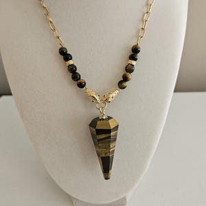 Op de afbeelding: Een goudkleurige ketting met een hanger. De hanger heeft een facetvormige, langwerpige vorm met bruine en gouden tijgeroogsteen. De ketting heeft tijgeroog kralen en decoratieve gouden accenten.