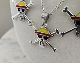 May include: Silver-toned necklaces featuring a skull and crossbones pendant. The pendant has a yellow hat with a red band, black eyes, and a silver skull. The chains are delicate and the pendants are detailed.