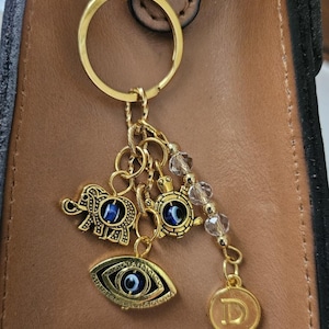 May include: Gold-toned keychain with charms. Charms include an elephant, turtle, and an eye, each with a blue center. A small gold disc with the letter "D" hangs from the chain. The keychain is attached to a brown leather surface.