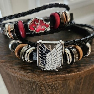 May include: Two black braided leather bracelets. One has a red cloud charm, red wooden beads, and silver accents. The other features a silver emblem with black and white wings, wooden beads, and silver accents. The bracelets are stacked.