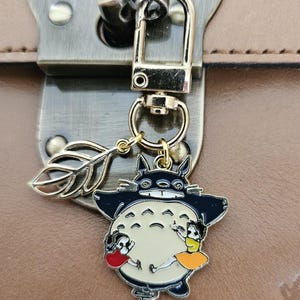 Puede incluir: Un llavero dorado con un personaje de dibujos animados con cuerpo negro y blanco, sosteniendo dos pequeñas figuras. El llavero está sujeto a un cierre metálico y un dije decorativo de hoja. El fondo es una superficie de cuero marrón.