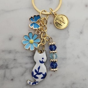 May include: A gold-colored keychain with a lobster clasp and a split ring. The keychain features a charm with a white cat design with blue floral accents, blue flower charms, and blue beaded accents. A gold-colored charm with the letter "W" is also included.