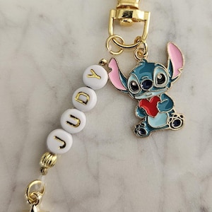 May include: A gold-colored keychain with a blue and pink cartoon character holding a red heart. The keychain has white beads spelling "JUDY" and a gold star charm. The character is a popular animated figure.
