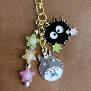 May include: A gold-colored keychain with multiple charms. Charms include a star, a black soot sprite, and a gray Totoro character. Additional charms are star-shaped and in shades of pink, yellow, and green.