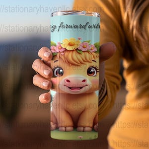 May include: A cylindrical tumbler featuring a cartoon cow with a floral crown. The tumbler has the text "g forward wit" on a light blue background. The cow is light brown with big eyes and a pink nose.