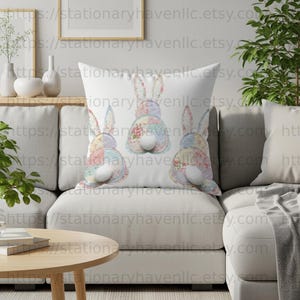 Pode incluir: Almofada decorativa branca com três desenhos de coelhos de patchwork. Cada coelho tem uma cauda branca fofa e corpos estampados com flores e cores pastel. A almofada está em um sofá branco, perfeita para a decoração de Páscoa.