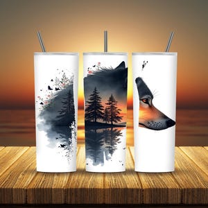 May include: Three white insulated tumblers with silver straws. Each tumbler features a different wolf-themed design: a forest scene, a wolf's head, and a forest with a sunset background. The tumblers are on a wooden surface.