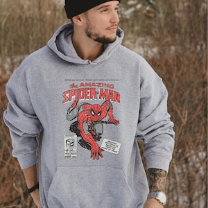 K&ouml;nnte beinhalten: Sportgrauer Hoodie mit einem Comic-Design von Spider-Man. Das Design enth&auml;lt den Text "the AMAZING SPIDER-MAN" in Rot und Wei&szlig;, zusammen mit zus&auml;tzlichem Text und Illustrationen. Der Hoodie hat eine K&auml;ngurutasche und eine Kapuze mit Kordelzug.