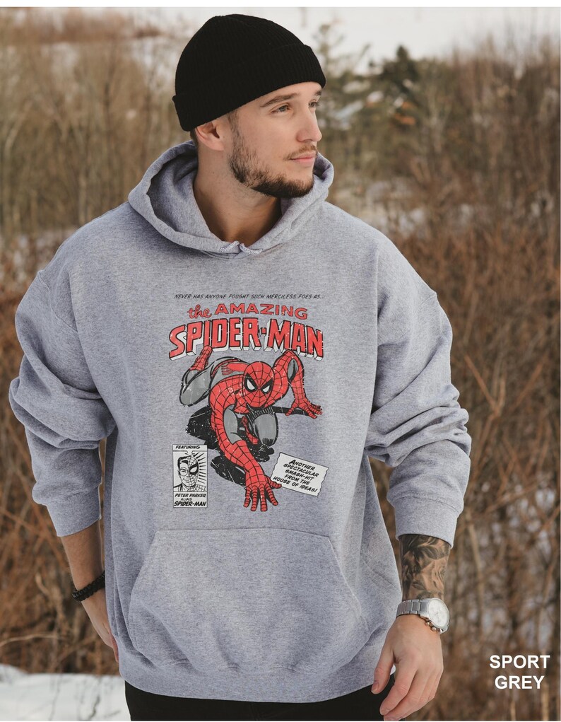 K&ouml;nnte beinhalten: Sportgrauer Hoodie mit einem Comic-Design von Spider-Man. Das Design enth&auml;lt den Text "the AMAZING SPIDER-MAN" in Rot und Wei&szlig;, zusammen mit zus&auml;tzlichem Text und Illustrationen. Der Hoodie hat eine K&auml;ngurutasche und eine Kapuze mit Kordelzug.