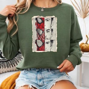 Op de afbeelding: Groen sweatshirt met een afbeelding van Spider-Mans masker en drie gezichten in een verweerde rechthoekige lijst. De sweatshirt is effen groen van kleur en heeft lange mouwen.