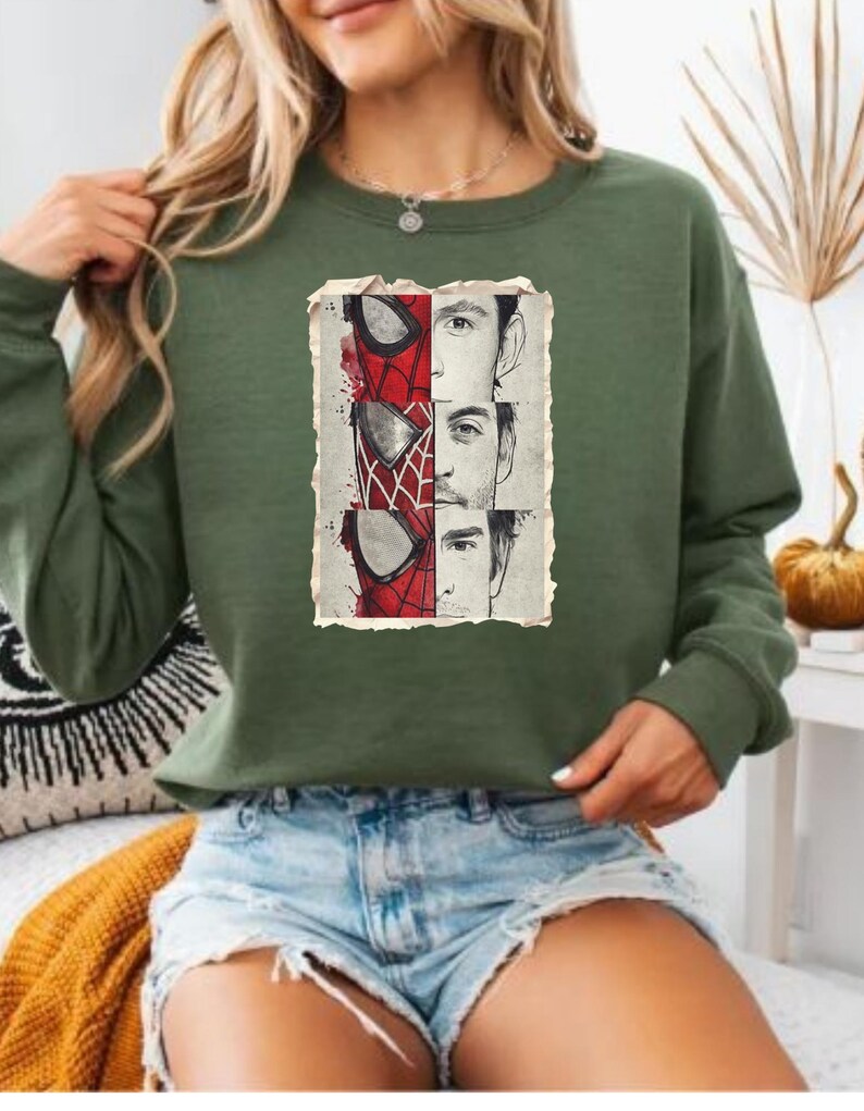 Op de afbeelding: Groen sweatshirt met een afbeelding van Spider-Mans masker en drie gezichten in een verweerde rechthoekige lijst. De sweatshirt is effen groen van kleur en heeft lange mouwen.