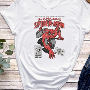 K&ouml;nnte beinhalten: Wei&szlig;es T-Shirt mit einer Comic-Grafik von Spider-Man in Rot und Grau, mit dem Text "the AMAZING SPIDER-MAN". Zus&auml;tzlicher Text enth&auml;lt "NEVER HAS ANYONE FOUGHT SUCH MERCILESS FOES AS..." und "ANOTHER SPECTACULAR SMASH-HIT FROM THE HOUSE OF IDEAS!"