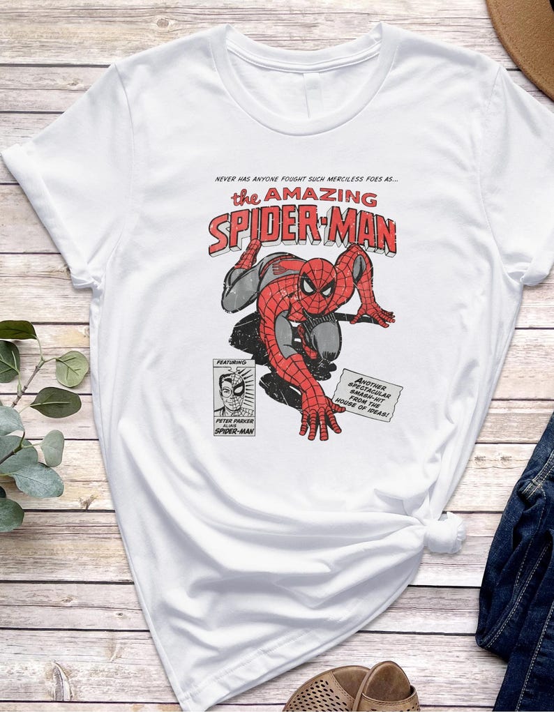 K&ouml;nnte beinhalten: Wei&szlig;es T-Shirt mit einer Comic-Grafik von Spider-Man in Rot und Grau, mit dem Text "the AMAZING SPIDER-MAN". Zus&auml;tzlicher Text enth&auml;lt "NEVER HAS ANYONE FOUGHT SUCH MERCILESS FOES AS..." und "ANOTHER SPECTACULAR SMASH-HIT FROM THE HOUSE OF IDEAS!"