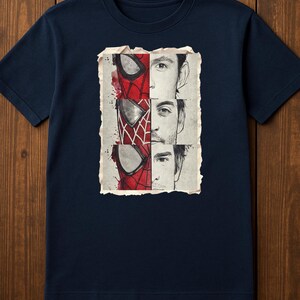 Op de afbeelding: Marineblauw T-shirt met een grafische afbeelding van Spider-Mans masker en gezicht. Het ontwerp is verdeeld in drie secties, elk met een andere Spider-Man-iteratie. De illustratie is geplaatst tegen een vintage achtergrond.