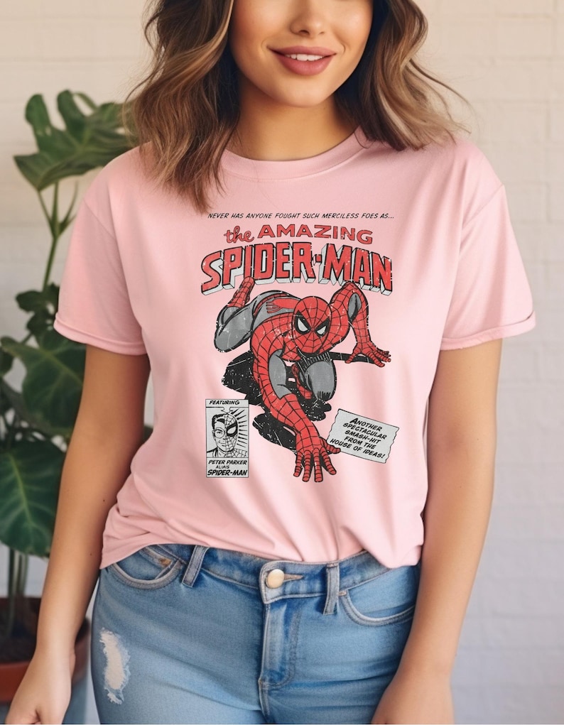 K&ouml;nnte beinhalten: Hellrosa T-Shirt mit einem Vintage-Comic-Design von Spider-Man. Die Grafik enth&auml;lt den Text "The Amazing Spider-Man" und andere Comic-Details. Das Shirt wird mit einer blauen Jeans kombiniert.