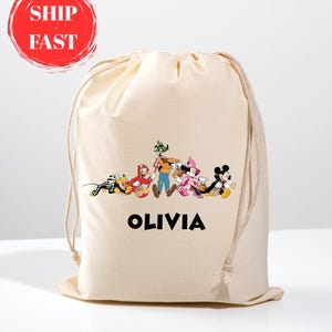 May include: A beige drawstring bag featuring a colorful graphic of Disney characters, including Mickey Mouse, Minnie Mouse, and Goofy. The name "OLIVIA" is printed in black below the characters. The bag has a simple, casual design.