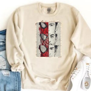 Op de afbeelding: Cr&egrave;mekleurige sweatshirt met een afbeelding van Spider-Mans masker en de gezichten van drie acteurs die het personage hebben gespeeld. De afbeelding is in rood, zwart en wit. Witte sneakers en een blauwe spijkerbroek zijn zichtbaar.
