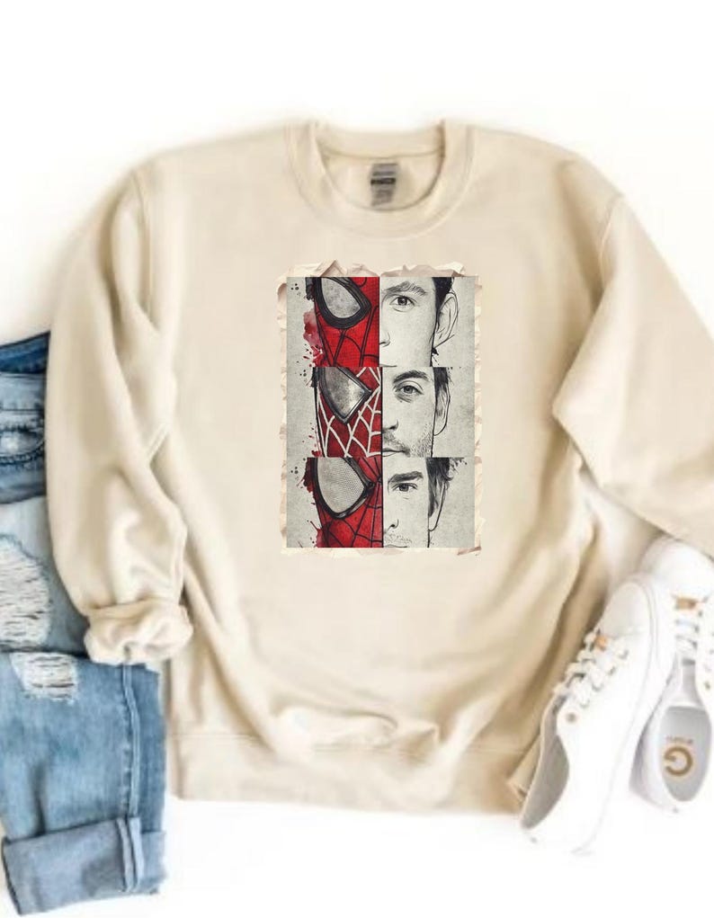 Op de afbeelding: Cr&egrave;mekleurige sweatshirt met een afbeelding van Spider-Mans masker en de gezichten van drie acteurs die het personage hebben gespeeld. De afbeelding is in rood, zwart en wit. Witte sneakers en een blauwe spijkerbroek zijn zichtbaar.