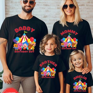K&ouml;nnte beinhalten: Schwarze T-Shirts f&uuml;r eine Familie, jedes mit einem Geburtstagsthema. Die Shirts zeigen ein Zirkuszelt-Design mit Cartoon-Figuren und den Texten "Daddy", "Mommy", "Brother" und "Amelia the Birthday Girl". Ballons und farbenfrohe Akzente vervollst&auml;ndigen das Design.