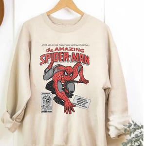 K&ouml;nnte beinhalten: Ein beiges Sweatshirt mit einer Comic-Grafik von Spider-Man in Rot, Schwarz und Grau. Der Text "the AMAZING SPIDER-MAN" ist deutlich sichtbar. Zus&auml;tzlicher Text lautet "NEVER HAS ANYONE FOUGHT SUCH MERCILESS FOES AS..." und "ANOTHER SPECTACULAR SMASH-HIT FROM THE HOUSE OF IDEAS!"