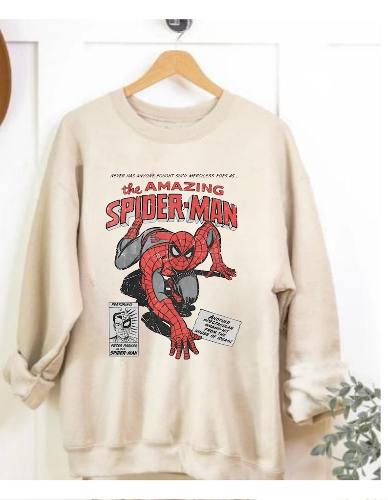 K&ouml;nnte beinhalten: Ein beiges Sweatshirt mit einer Comic-Grafik von Spider-Man in Rot, Schwarz und Grau. Der Text "the AMAZING SPIDER-MAN" ist deutlich sichtbar. Zus&auml;tzlicher Text lautet "NEVER HAS ANYONE FOUGHT SUCH MERCILESS FOES AS..." und "ANOTHER SPECTACULAR SMASH-HIT FROM THE HOUSE OF IDEAS!"